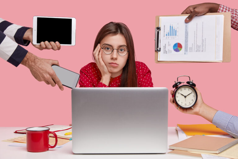 Serious Caucasian woman with dark hair, wears round spectacles, red shirt, sits in front of laptop computer drinks beverage, unrecognizable people hold alarmclock, papers, touchpad, cellular in hands