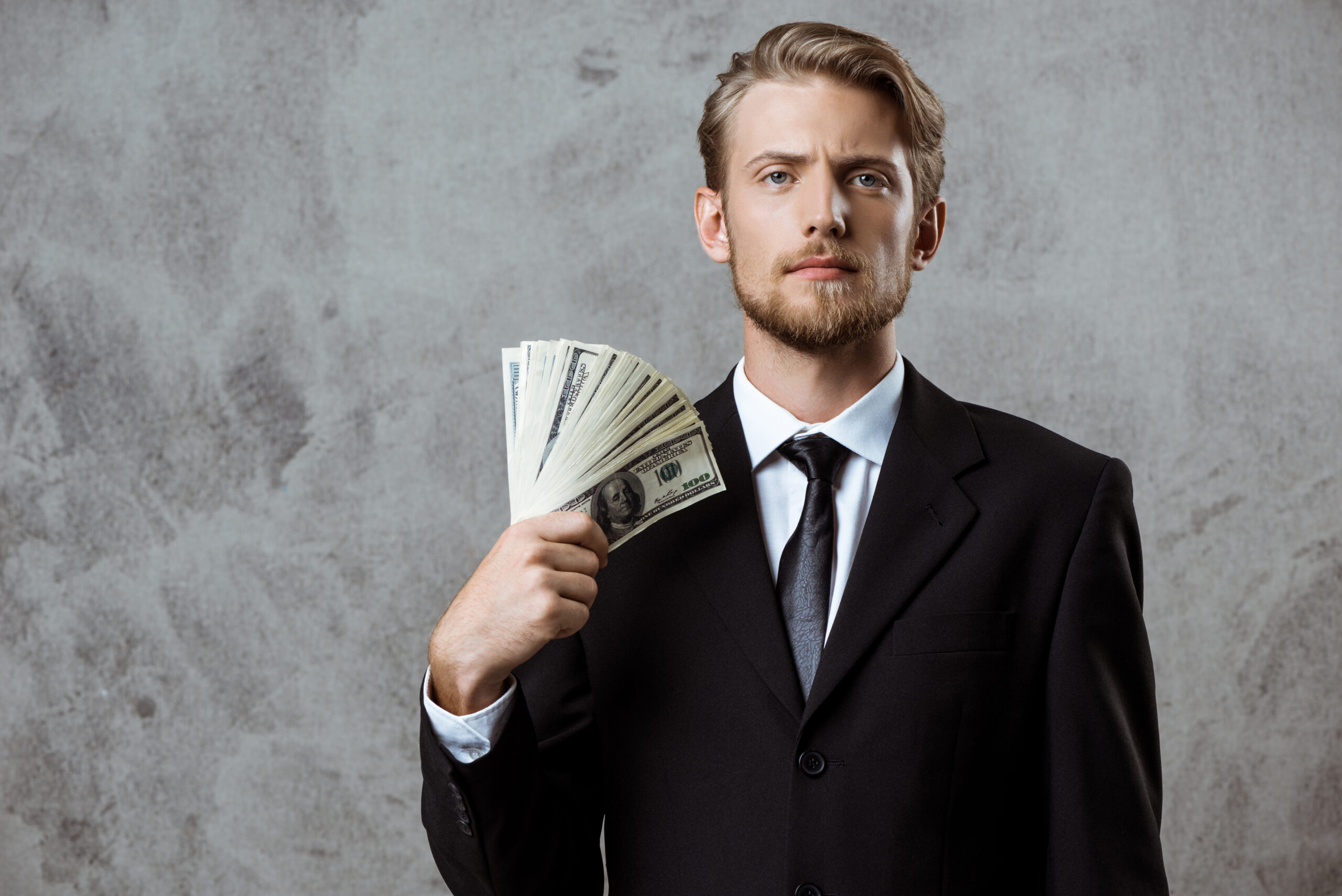 Young successful businessman in suit holding money over grey background.